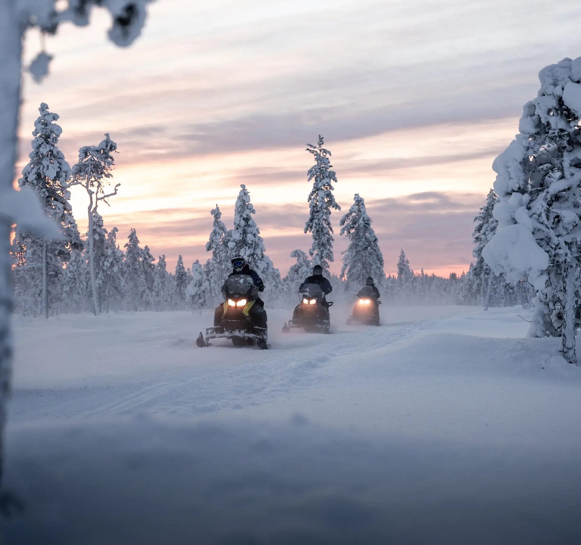 Tres motos de nieve recorriendo un bosque nevado al atardecer