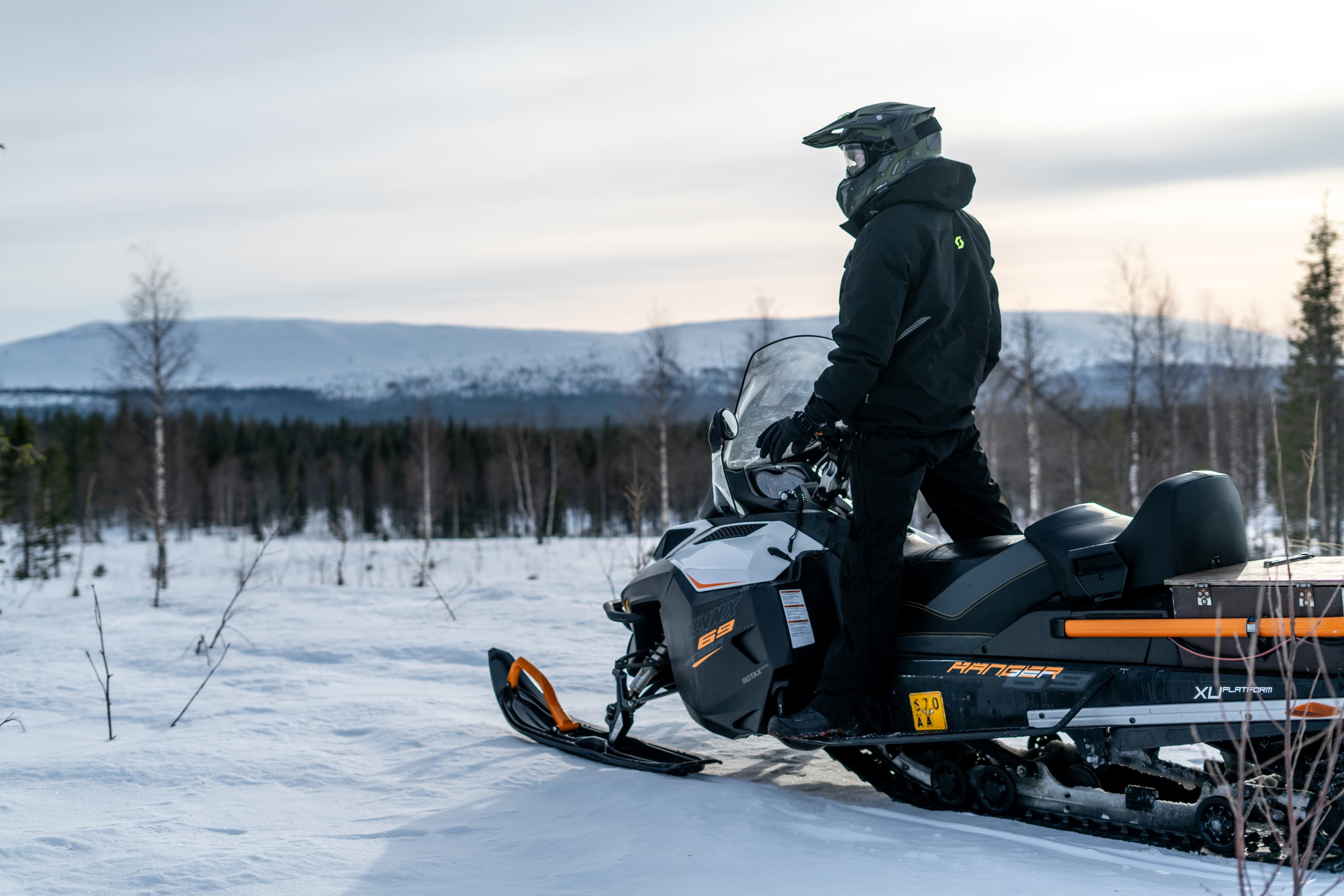 Una persona de pie sobre una moto de nieve al anochecer, con colinas al fondo